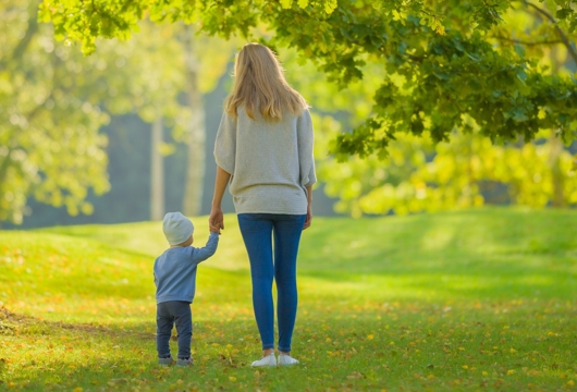 Young adult mother and baby boy standing on grass at city park and looking far away. Spending time together in beautiful warm sunny autumn day. Back view.