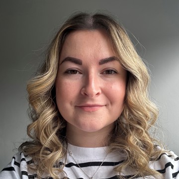  Headshot of Molly, a woman with shoulder-length curly blonde hair, wearing a black and white striped top and a silver necklace, smiling at the camera against a grey background.