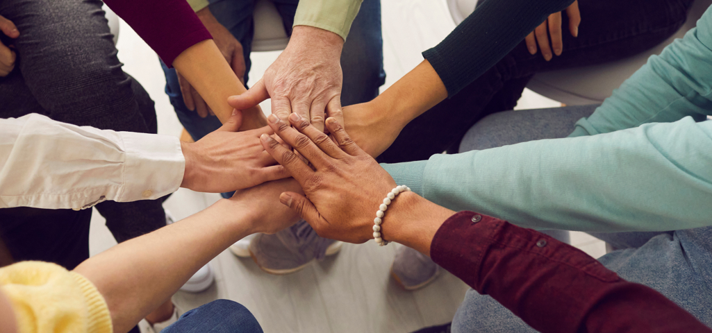 Seven people's hands all on top of each other in the middle to represent a team