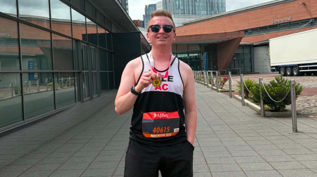 Liam stands on a paved walkway between modern buildings, wearing running gear, holding the medal up toward the camera under a cloudy sky.