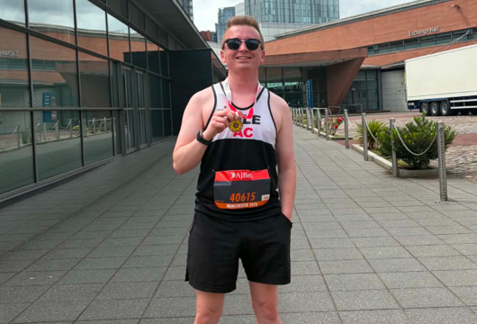 Liam stands on a paved walkway between modern buildings, wearing running gear, holding the medal up toward the camera under a cloudy sky.