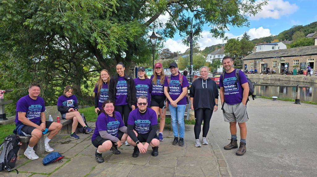 A group of colleagues wearing purple Candlelighters charity walk T-shirts smile for a photo beside a canal in Yorkshire during their 50km fundraising walk.