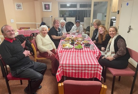 A photograph inside a community centre with people having a roast dinner and smiling. 