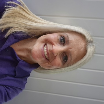 A person with straight, shoulder‑length blonde hair is wearing a purple Yorkshire Housing polo shirt. They are stood in front of a light grey paneled wall taking a selfie-style photograph.