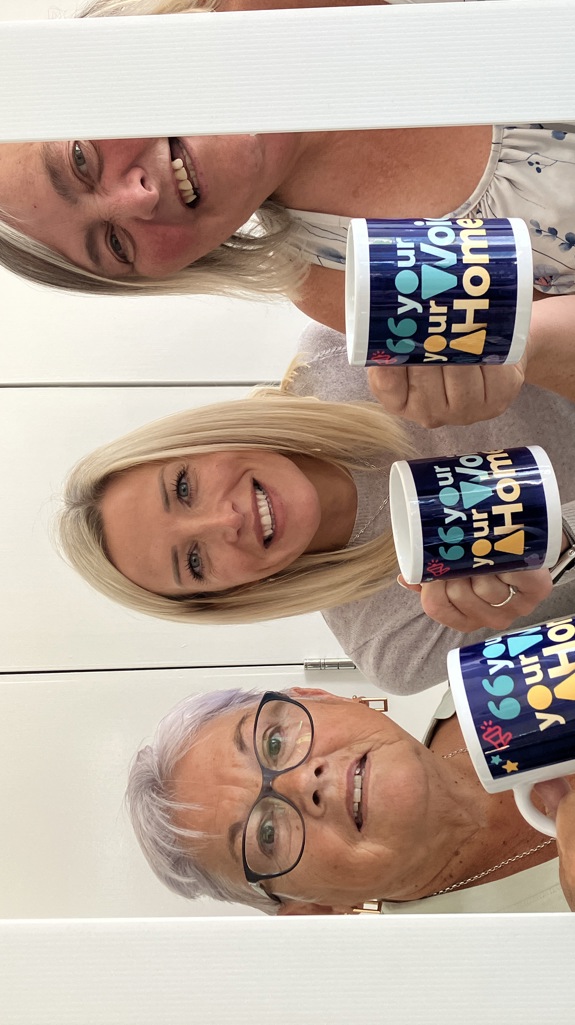 Three women smiling holding mugs with colourful ‘Your Voice Your Home’ designs, standing behind a large Instagram-style photo frame labelled Yorkshire Housing CommuniTEA.