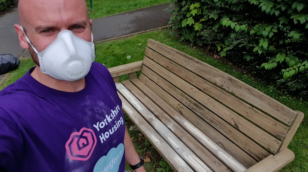  Our Executive Director of Finance and Governance, Rob, wearing a white dust mask and a purple Yorkshire Housing t-shirt sanding down a wooden bench outside on a grassy area.