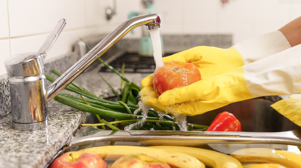 Photo of someone washing a tomato under the tap with spring onions in the sink and apples and bananas in the water at the bottom of the photo. 