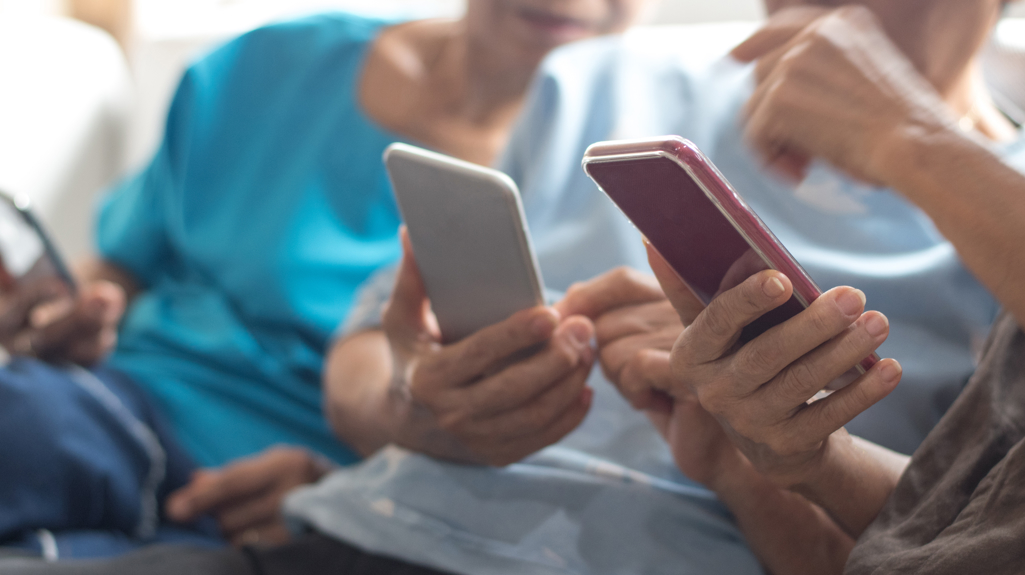 Three people sat on a sofa holding phones, helping each other use them. 