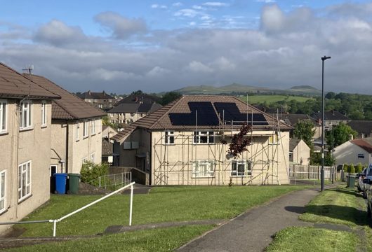 A photograph of three houses, the house in the centre of the image is having solar panels installed. It's a cloudy but blue skied day.