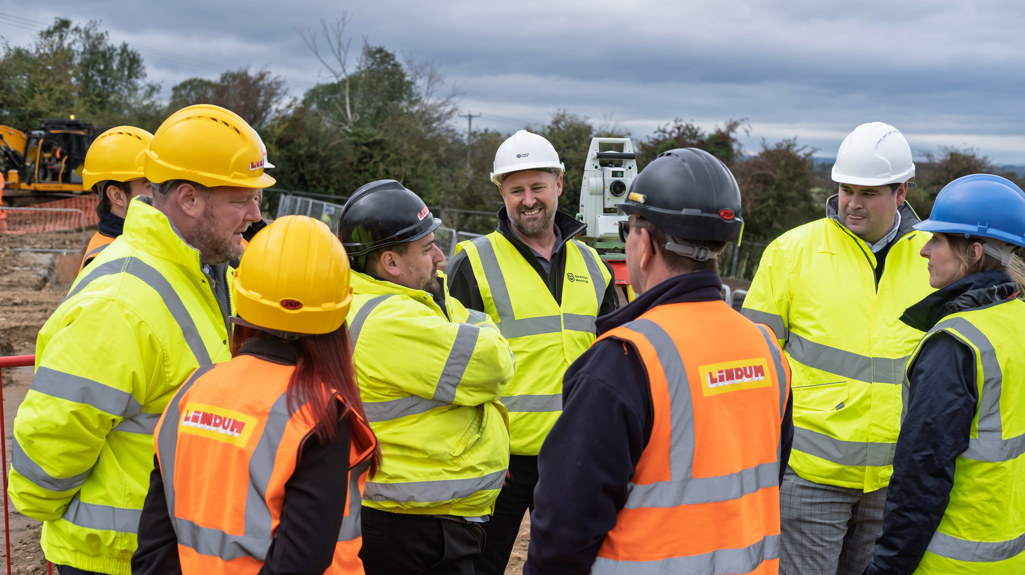 A small group of people in hard hats and high-visibility jackets gather in discussion at a construction site. One man at the centre is smiling as he speaks to others, while heavy machinery and fencing can be seen behind them.