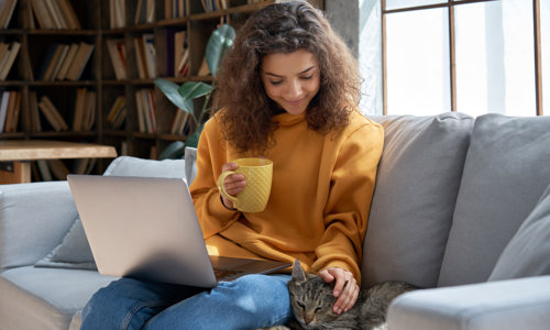 Women sitting on the sofa in a yellow jumper, stroking a cat with a laptop on her knee