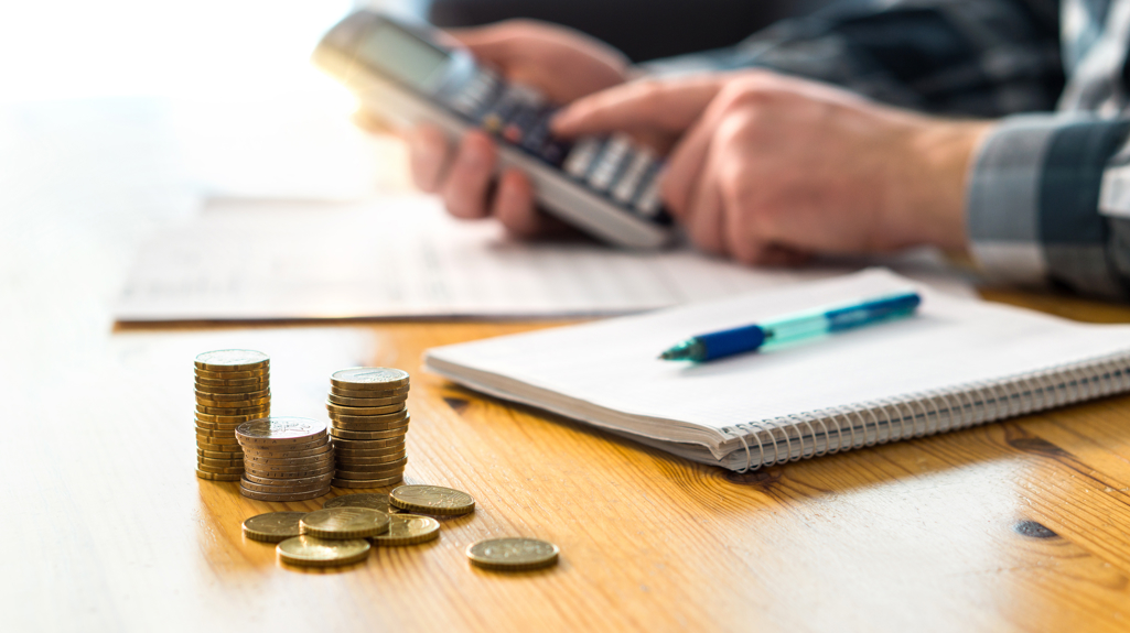 Man using a calculator at a table with a notepad and pen and a pile of coins
