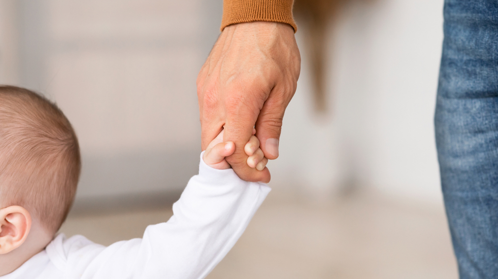 The side of a baby's head and arm holding an adult's hand