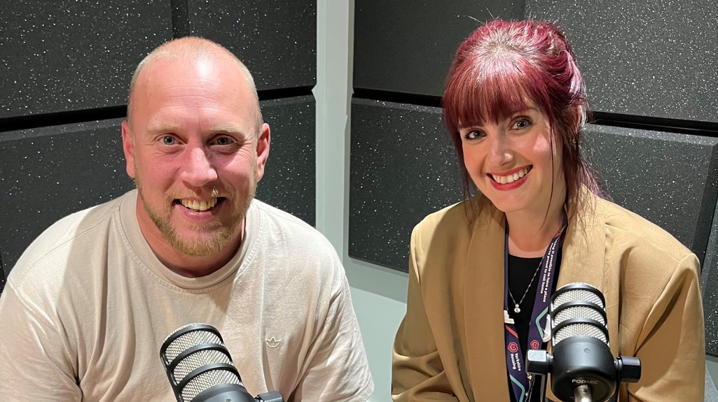 Joe, a man with a trimmed beard, wearing a light beige T-shirt, and Sian, a woman with dark red hair tied back, wearing a tan blazer over a black top with leopard-print detail on the sleeves, sit side by side in a recording studio, smiling at the camera with microphones in front of them.