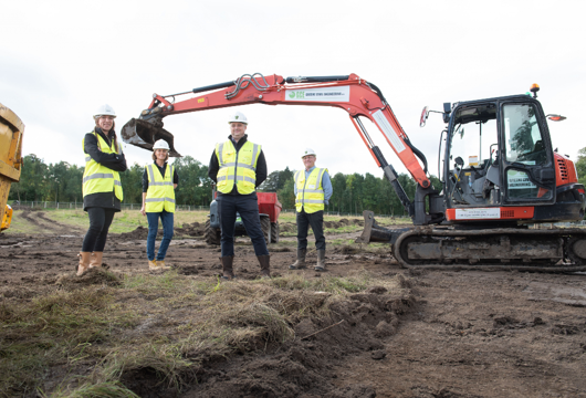 Four people stood on a development site in its first stages with a digger behind them. 