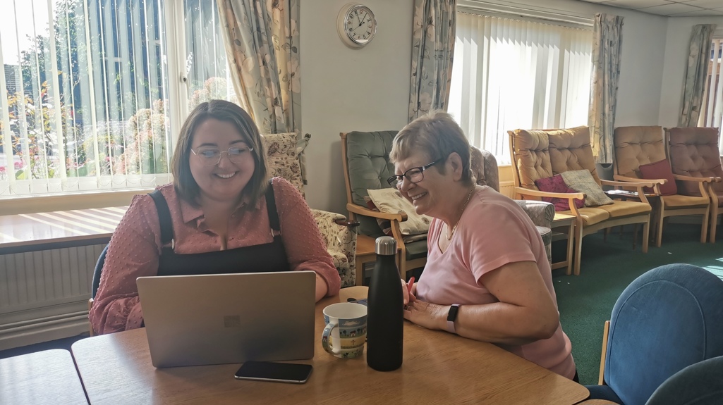 Two women sitting at a table in a cosy living room smiling and laughing together with a laptop, phone and a cup of tea at the table. 
