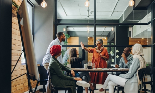 A group of colleagues celebrating with two women high fiving in the middle, in an office environment. 