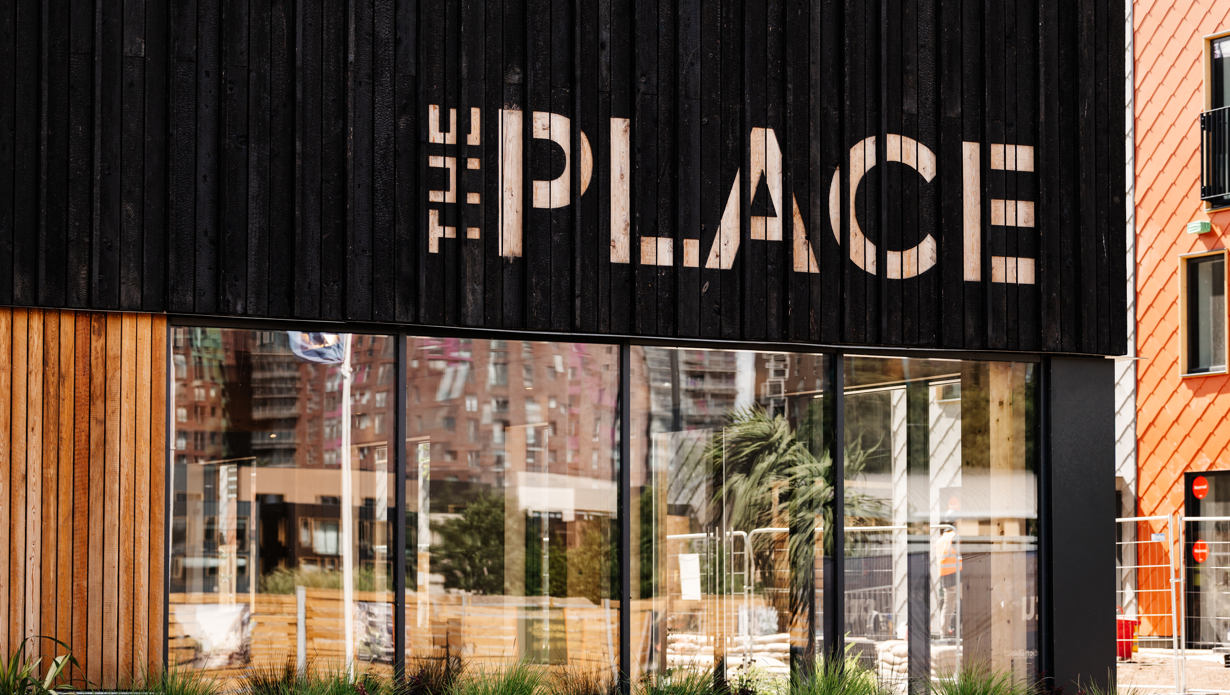 Photograph of the outside of The Place, showing a large glass wall with black wood above it with The Place spelled out in pale wood. 