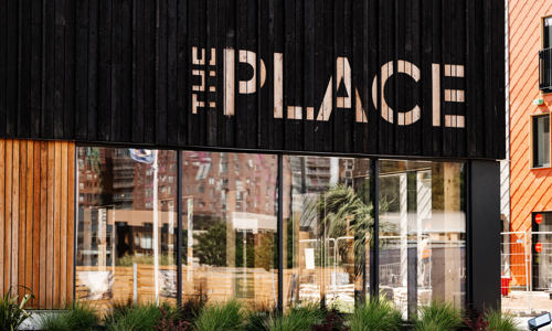 Photograph of the outside of The Place, showing a large glass wall with black wood above it with The Place spelled out in pale wood. 