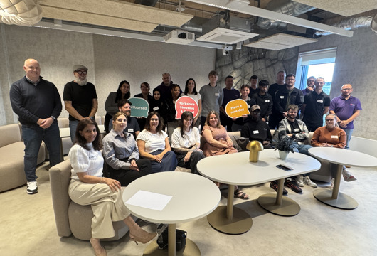 A group of Yorkshire Housing apprentices and their mentors pose together in a modern office lounge. Around twenty people of mixed ages and backgrounds stand and sit around small round tables, smiling at the camera. Some hold colourful speech-bubble signs, including one that says “Yorkshire Housing” and another that says “Have a nice day.” The space features grey walls, exposed ceiling ducts, and soft seating, creating a relaxed, contemporary setting.