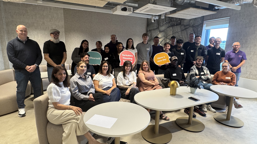 A group of Yorkshire Housing apprentices and their mentors pose together in a modern office lounge. Around twenty people of mixed ages and backgrounds stand and sit around small round tables, smiling at the camera. Some hold colourful speech-bubble signs, including one that says “Yorkshire Housing” and another that says “Have a nice day.” The space features grey walls, exposed ceiling ducts, and soft seating, creating a relaxed, contemporary setting.