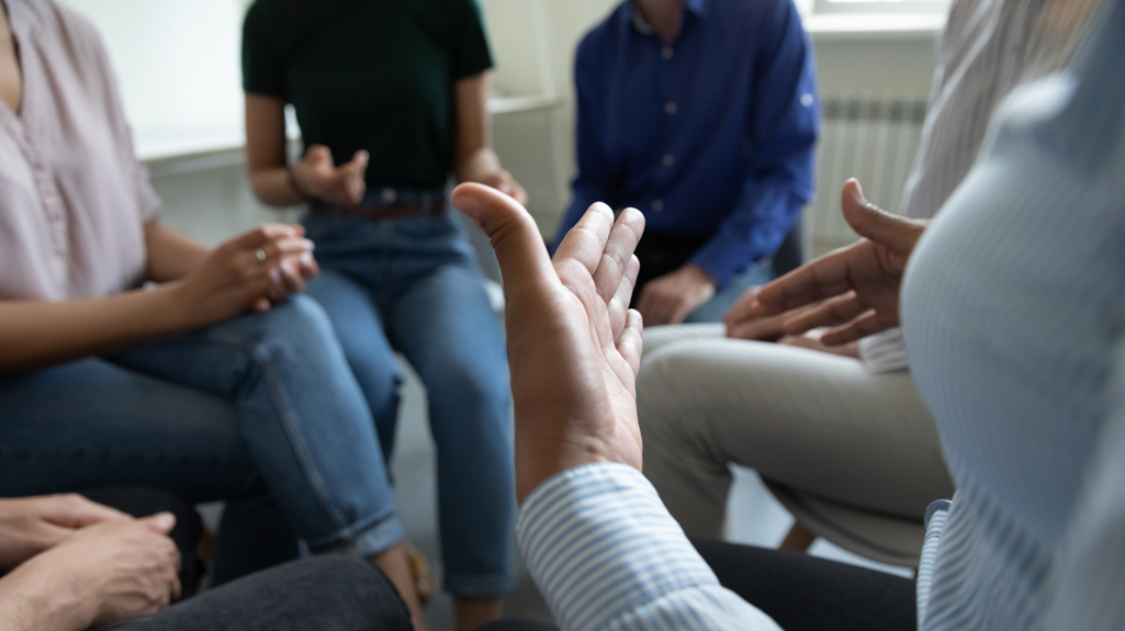 Abstract picture of people sat in a circle with one person gesturing with their hands
