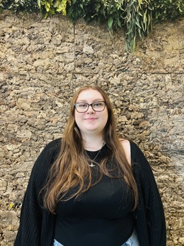 Photo of Amy our housing apprentice stood against a cork wall smiling