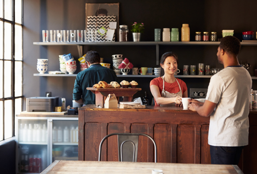 A photograph of a coffee shop with someone behind the counter smiling while serving a customer. 