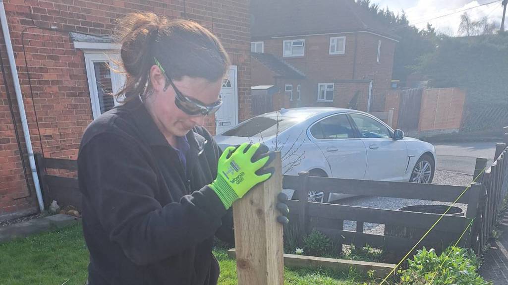 Person wearing safety glasses and gloves installing a wooden fence post into the ground beside a pavement, with string lines marking the fence line.