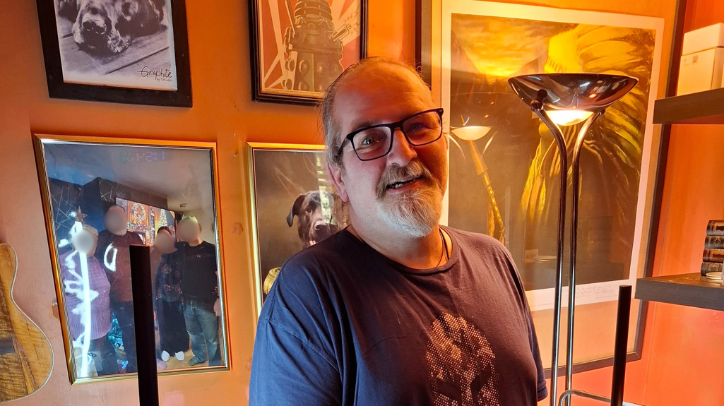 A photo of Rob standing and smiling at the camera in front of a wall of photographs 
