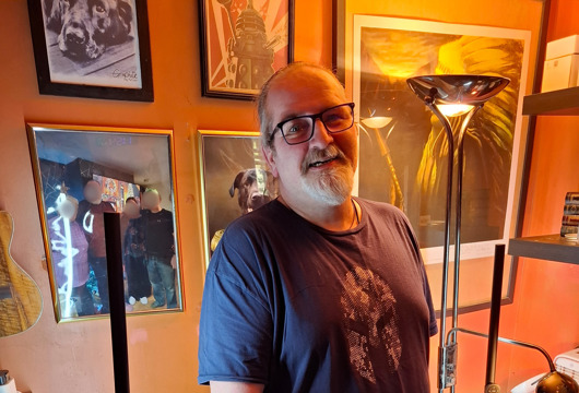 A photo of Rob standing and smiling at the camera in front of a wall of photographs