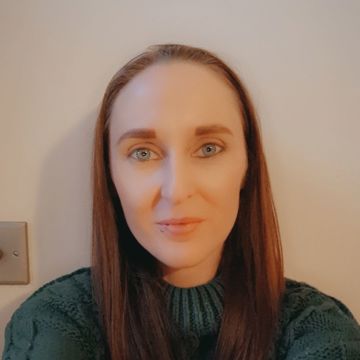 A person with long, straight brown hair is sitting against a plain light-colored wall. They are wearing a chunky, dark green knitted sweater, and their arms are slightly extended forward as if taking a selfie. 