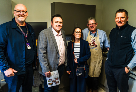 Five adults stand side by side in a small kitchen area, facing the camera. Some wear business attire while one person wears an apron. Kitchen cabinets and worktops are visible behind them, and one person holds a leaflet or booklet.