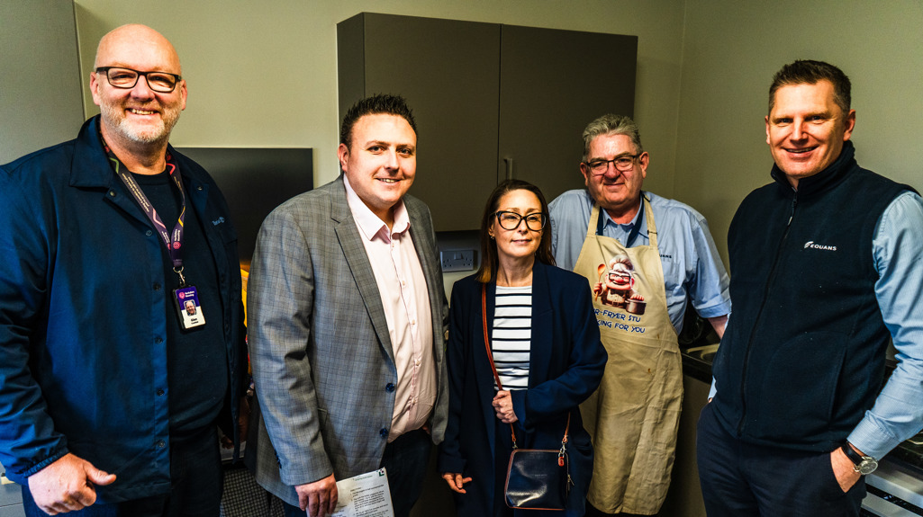 Five adults stand side by side in a small kitchen area, facing the camera. Some wear business attire while one person wears an apron. Kitchen cabinets and worktops are visible behind them, and one person holds a leaflet or booklet.