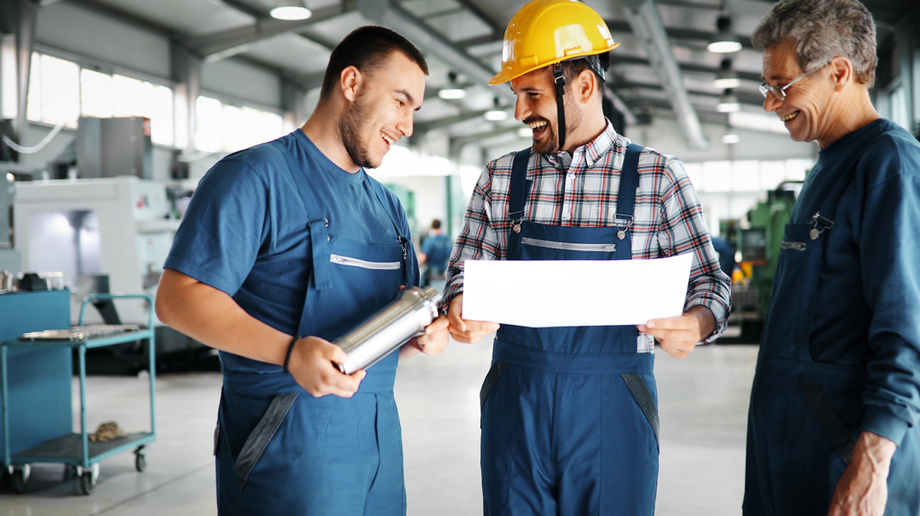 Three men stood in a warehouse or factory with overalls laughing together holding some paper and a metal tube.