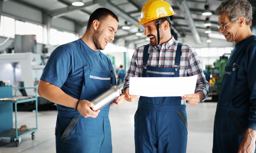 Three men stood in a warehouse or factory with overalls laughing together holding some paper and a metal tube.