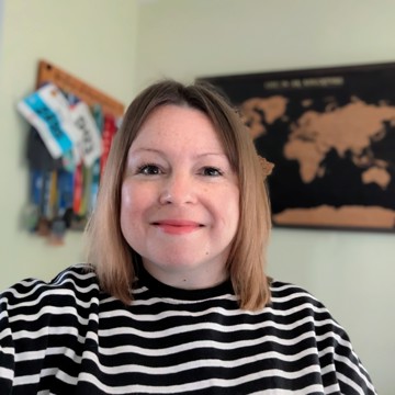 Victoria wearing a black‑and‑white striped jumper is indoors, with a world map poster on the wall behind her and a rack of colourful medals hanging to the left.