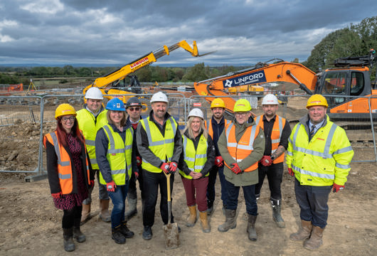 A group of construction workers and project representatives wearing high-visibility jackets, hard hats, and boots stand together at a muddy building site. They are smiling for a group photo with diggers and heavy machinery in the background under a cloudy sky.