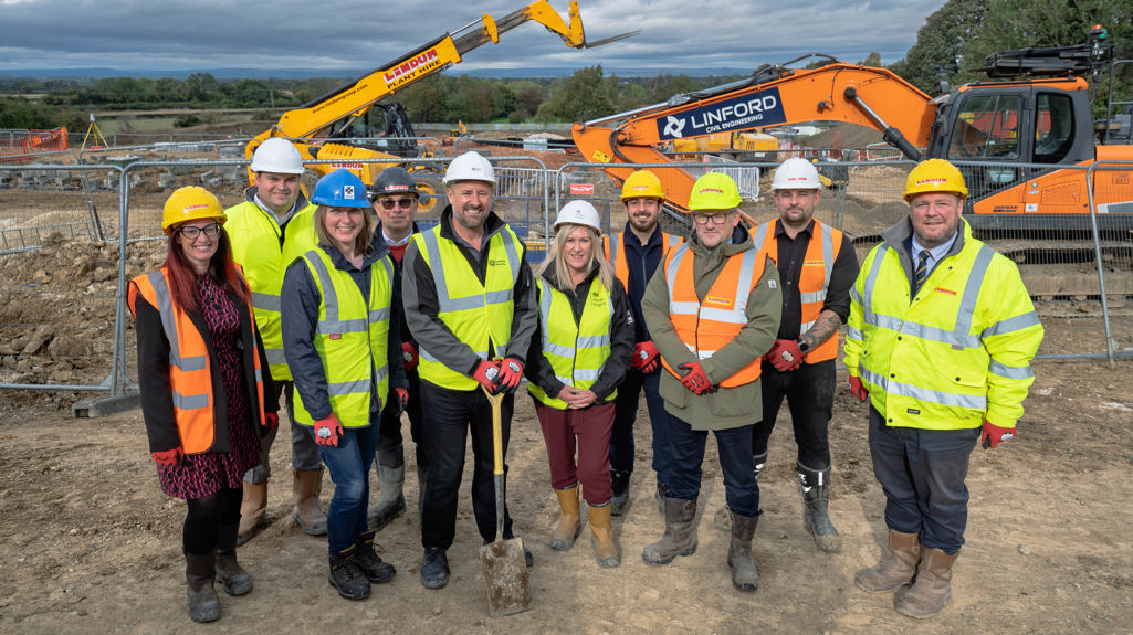 A group of construction workers and project representatives wearing high-visibility jackets, hard hats, and boots stand together at a muddy building site. They are smiling for a group photo with diggers and heavy machinery in the background under a cloudy sky.