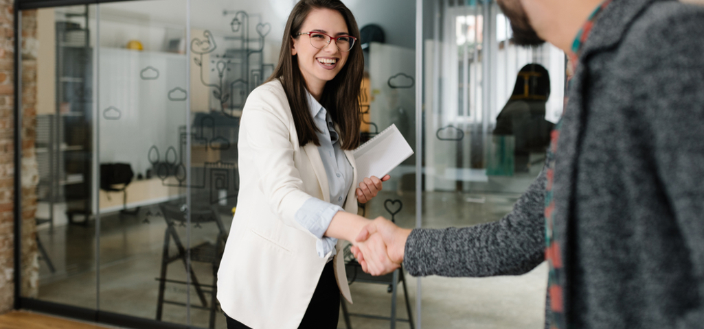 Woman in a white blazer and light blue shirt shaking the hand of a man wearing a more casual grey top. Both are smiling in a modern office.