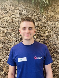 Photo of Jake our multi-skilled apprentice wearing a purple Yorkshire Housing t-shirt stood infront of a cork wall