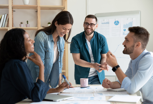 Four people in a work environment stood and sat round a table smiling an laughing