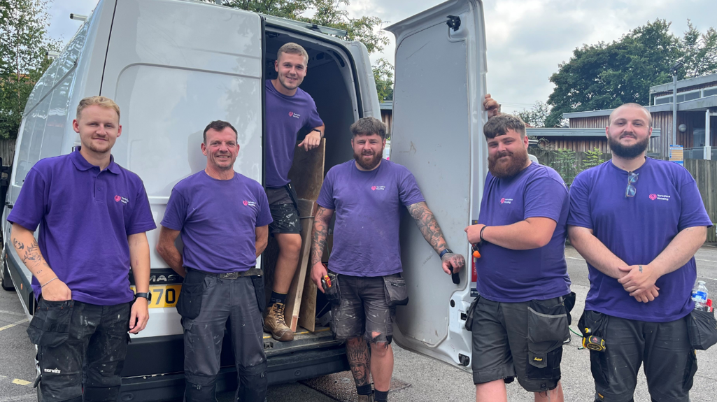 Six men in purple Yorkshire Housing t-shirts standing by the back of a white van, smiling at the camera, with tools and work trousers showing they are part of a maintenance team.