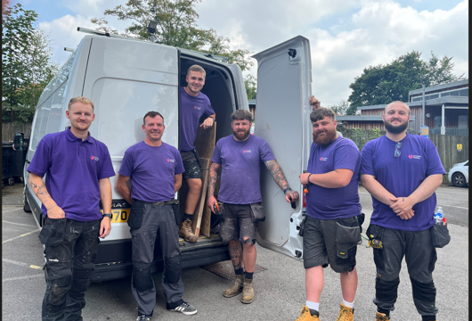 Six men in purple Yorkshire Housing t-shirts standing by the back of a white van, smiling at the camera, with tools and work trousers showing they are part of a maintenance team.