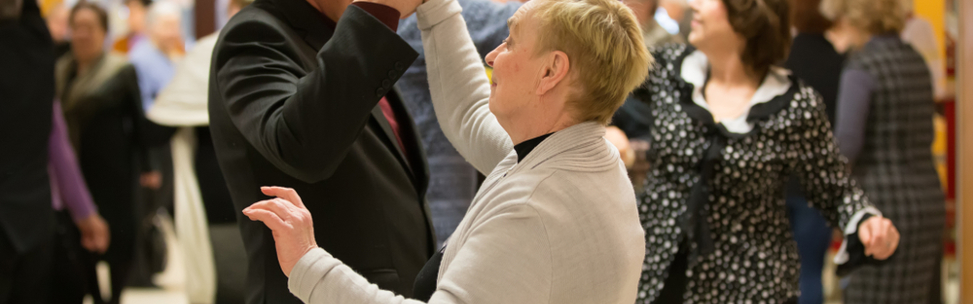 Two older people dancing together in a room where others are dancing too. 