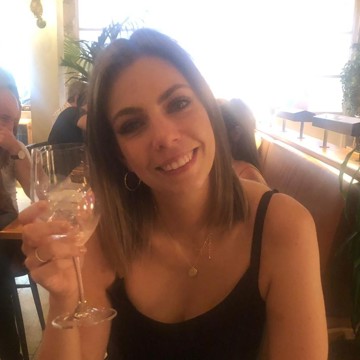 Laura smiling at the camera in a restaurant, holding up a glass with ice. She has straight, shoulder‑length light brown hair and is wearing a black sleeveless top, gold hoop earrings, and a gold necklace.
