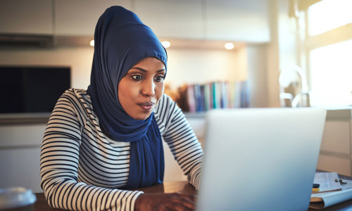 Women working at home, typing on a laptop while wearing a navy hijab and navy and white striped top. 