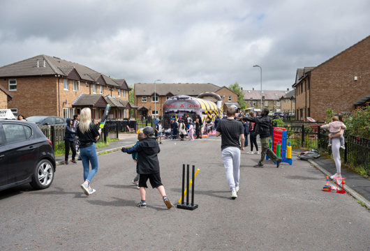 A number of people playing various street games like cricket and an obstacle course 