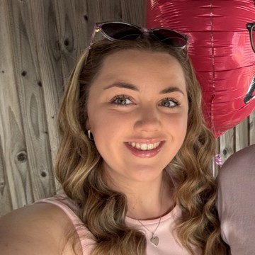 A woman with wavy, shoulder-length blonde hair is wearing a light pink sleeveless top and a heart-shaped necklace. She is standing outdoors in front of a wooden fence, with a large pink balloon beside her.