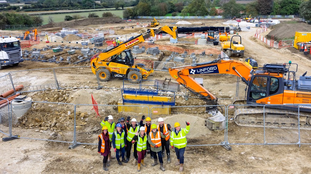 An aerial view of a busy construction site showing excavators, JCBs, and groundwork in progress. A group of people in high-visibility jackets and hard hats stand at the front of the site, waving towards the camera. The scene includes safety fencing, machinery, and building materials spread across the area.